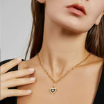A woman in a black V-neck top with her hand resting on her bare shoulder, modeling a gold paperclip chain lariat necklace that drops into a black enamel heart pendant.