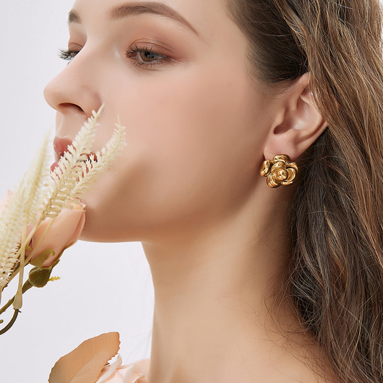 Close-up of the lower half of a woman's face and neck, holding pale pink roses, highlighting a gold camellia flower earring on her ear.