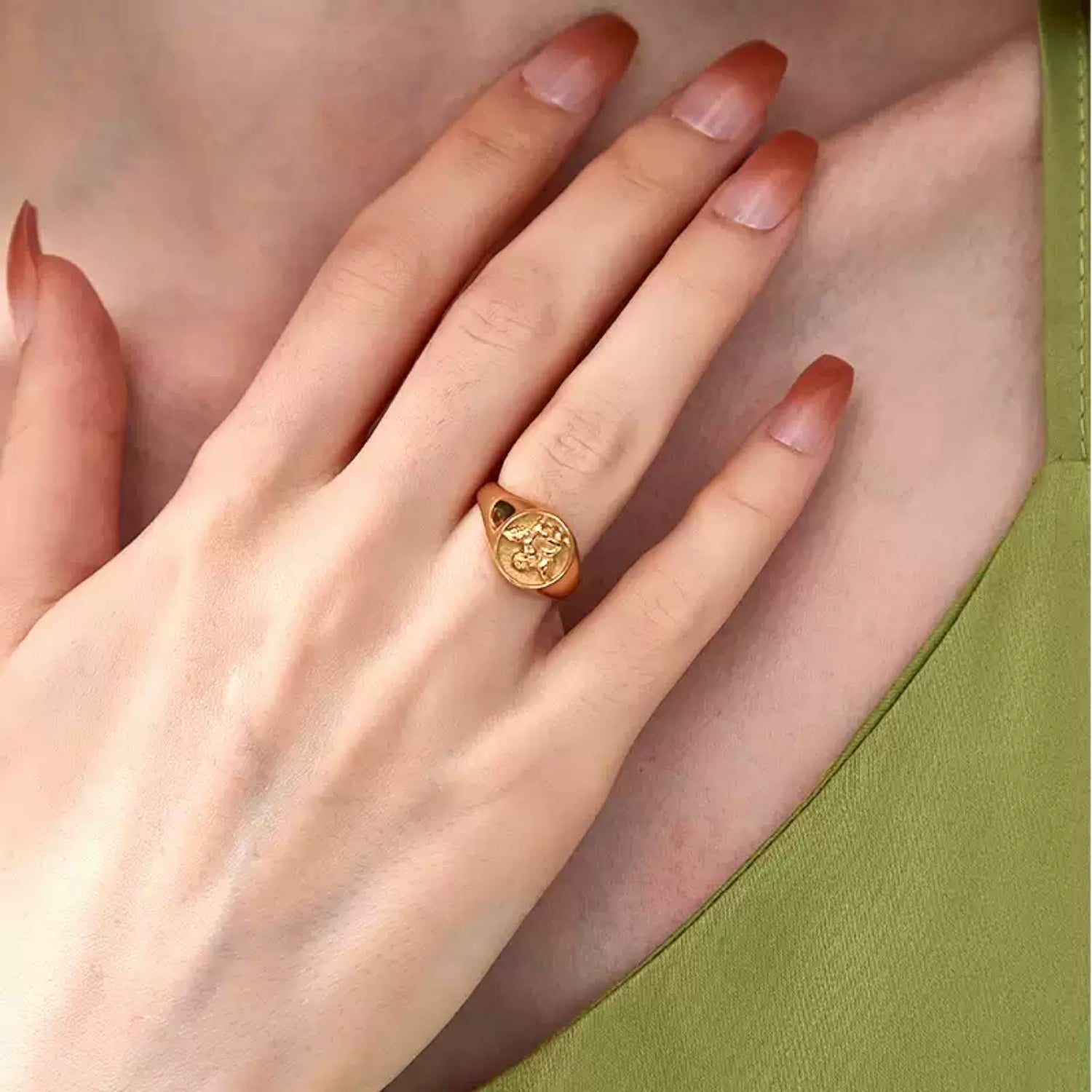 A woman's hand resting gracefully near her collarbone wearing a green silk top, showcasing a gold vintage angel signet ring on her middle finger.