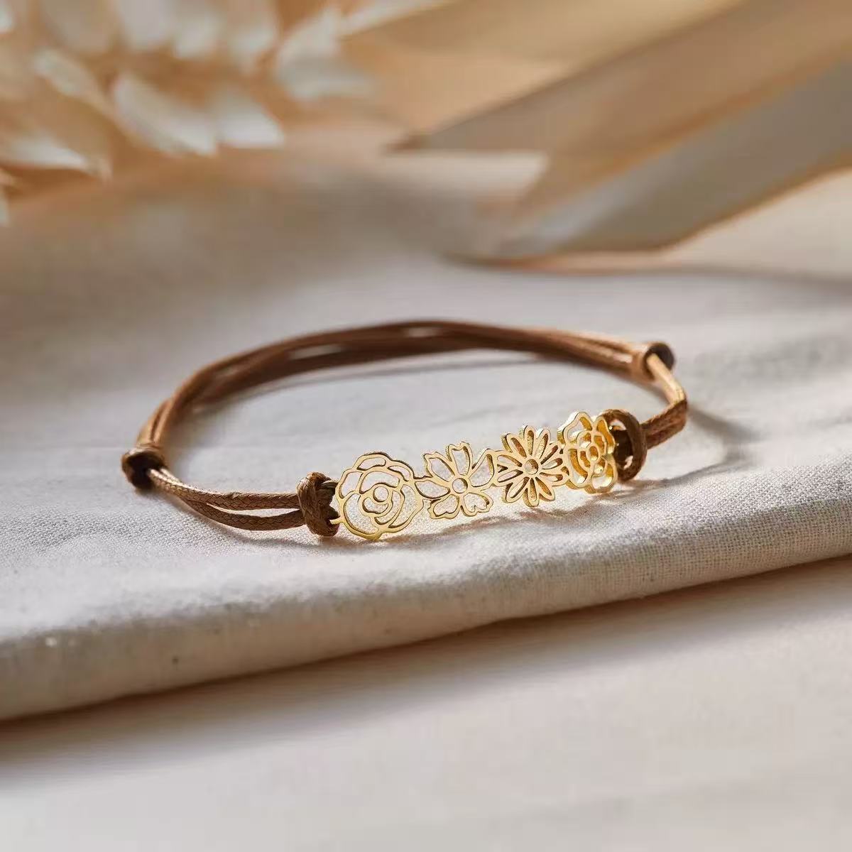 Close-up of a custom gold birth flower bracelet featuring three intricate cutout blooms on an earthy brown waxed cord.