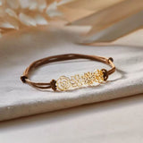Close-up of a custom gold birth flower bracelet featuring three intricate cutout blooms on an earthy brown waxed cord.