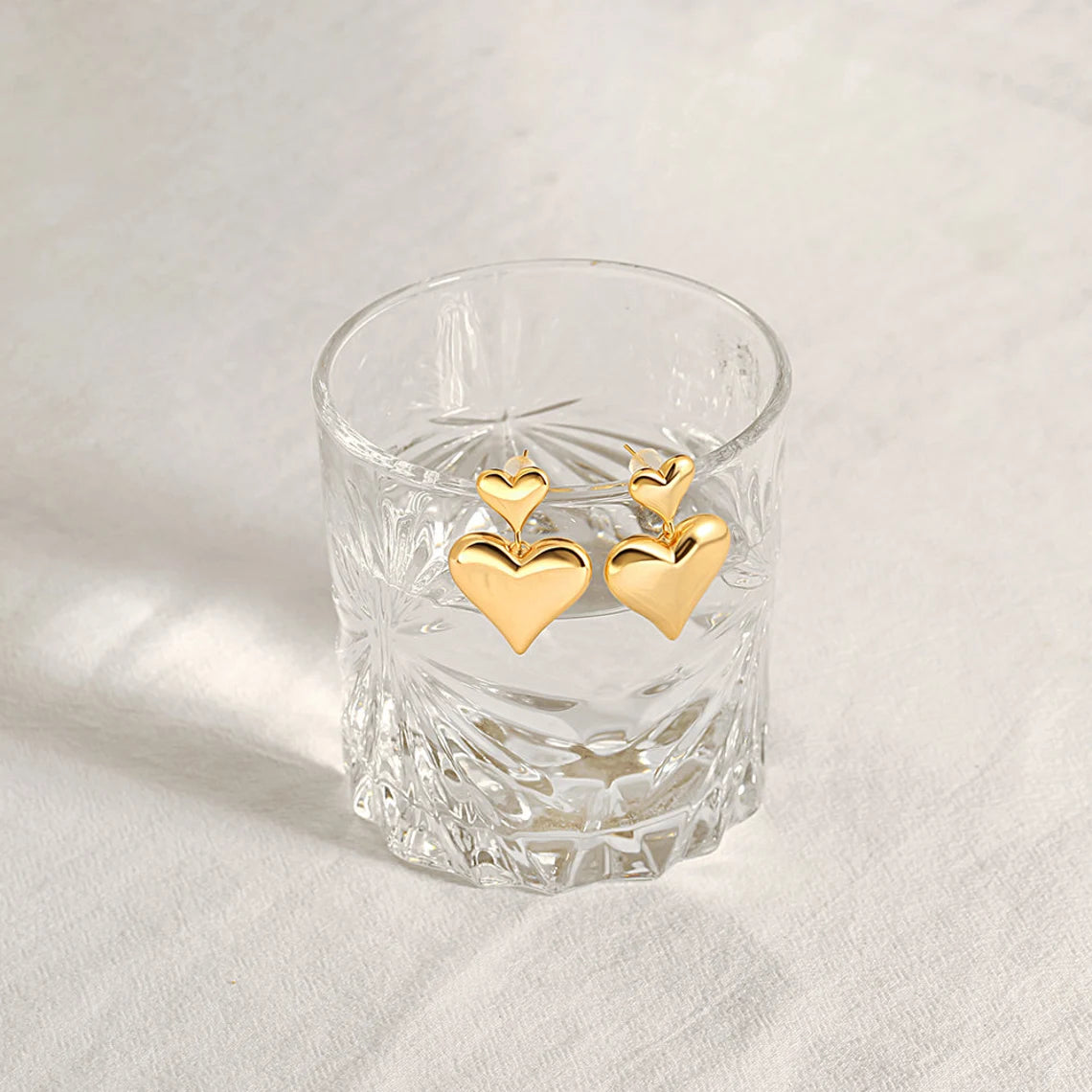 Two gold plated double puffy heart dangle earrings elegantly hanging on the rim of a clear textured glass.