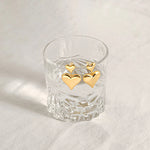 Two gold plated double puffy heart dangle earrings elegantly hanging on the rim of a clear textured glass.