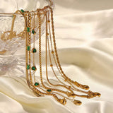 A collection of various gold necklaces and bracelets, including a snake chain with green baguette stones and chains with green heart pendants, draped elegantly over a clear crystal bowl on white silky fabric.