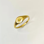 Angled view of a minimalist gold signet ring showcasing its polished surface and an engraved starburst pattern, resting on a white surface.