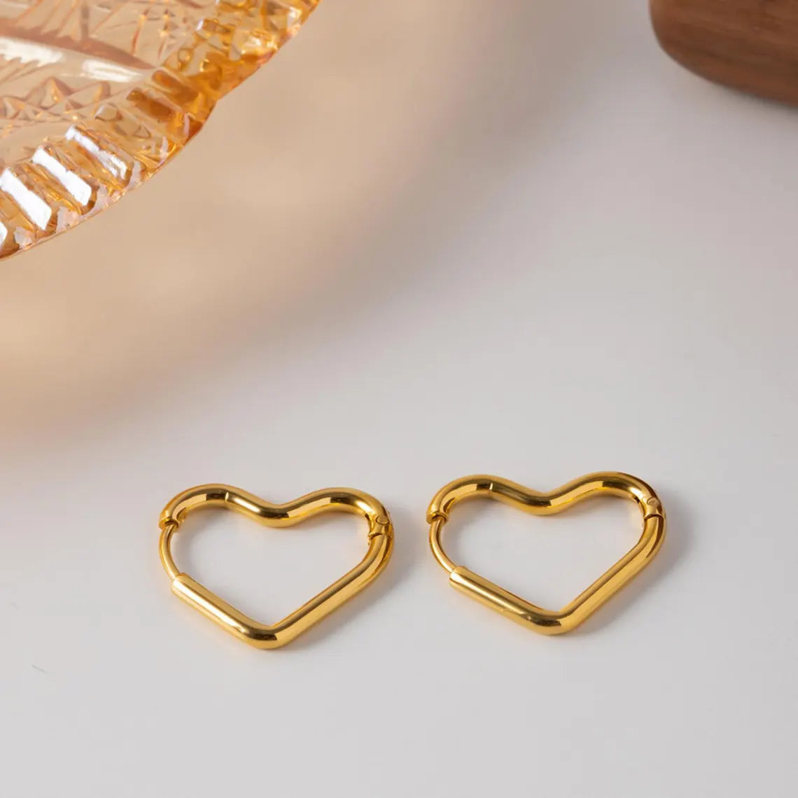 A pair of geometric gold heart hoop earrings placed on a clean white surface next to a textured amber glass tray.