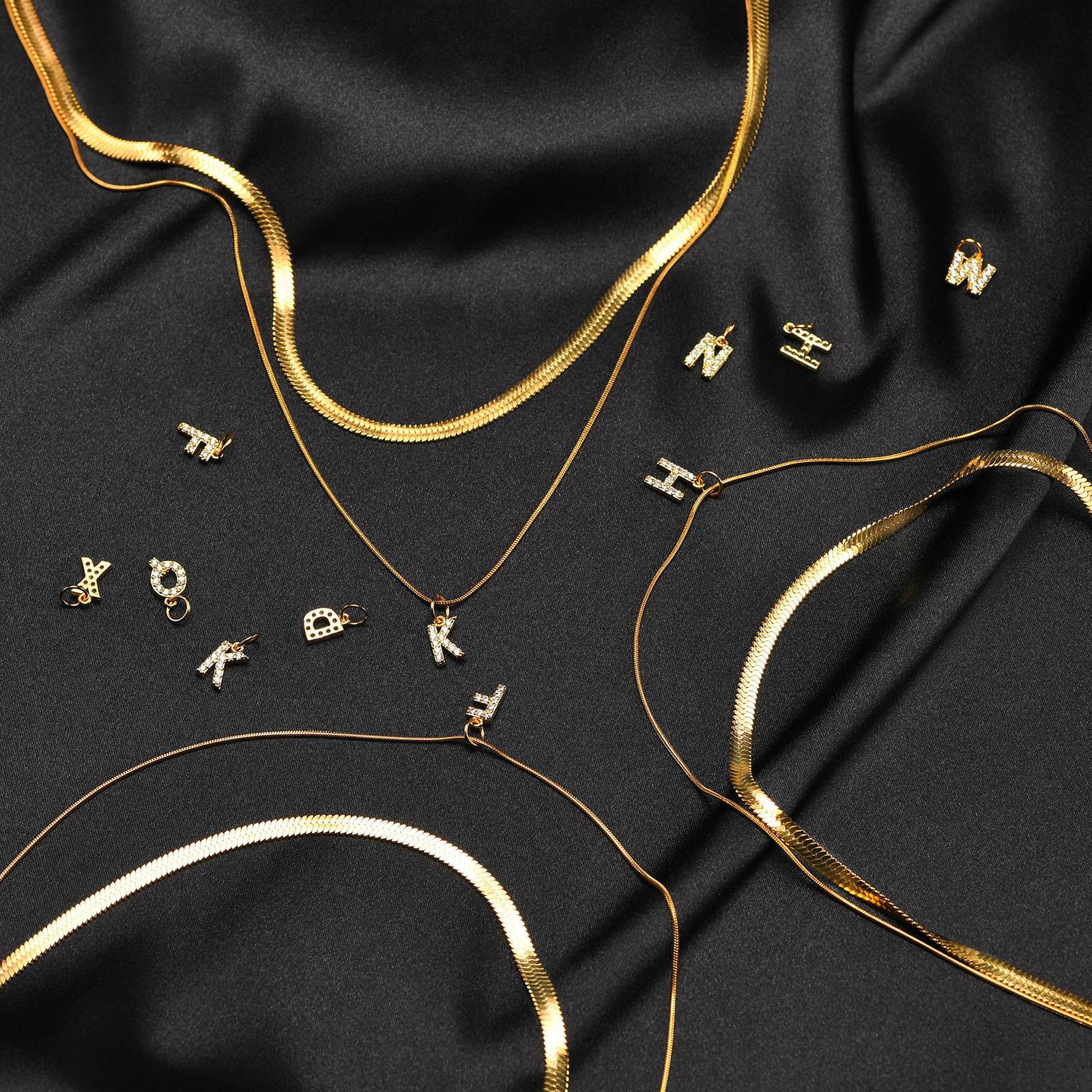 Flat lay of multiple gold herringbone snake chains and various sparkling pave cubic zirconia initial letter pendants scattered on black satin fabric.