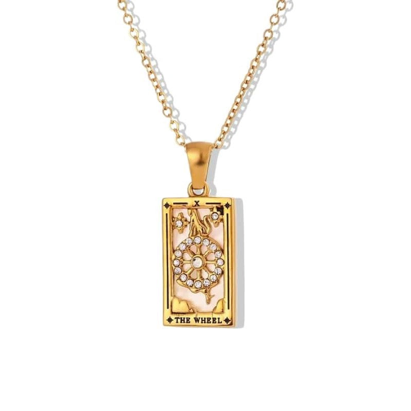 A gold rectangular tarot card pendant labeled THE WHEEL, featuring a white or pale pink enamel background with a gold wheel motif decorated with clear stones and a figure standing on top.