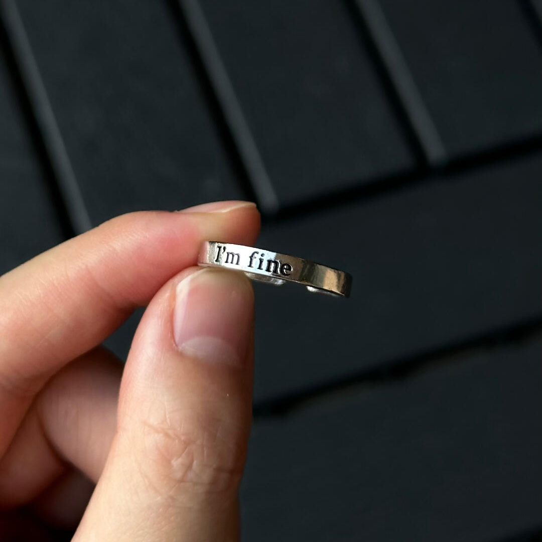 Fingers holding a delicate silver ring, clearly displaying the external engraved message "I'm fine."