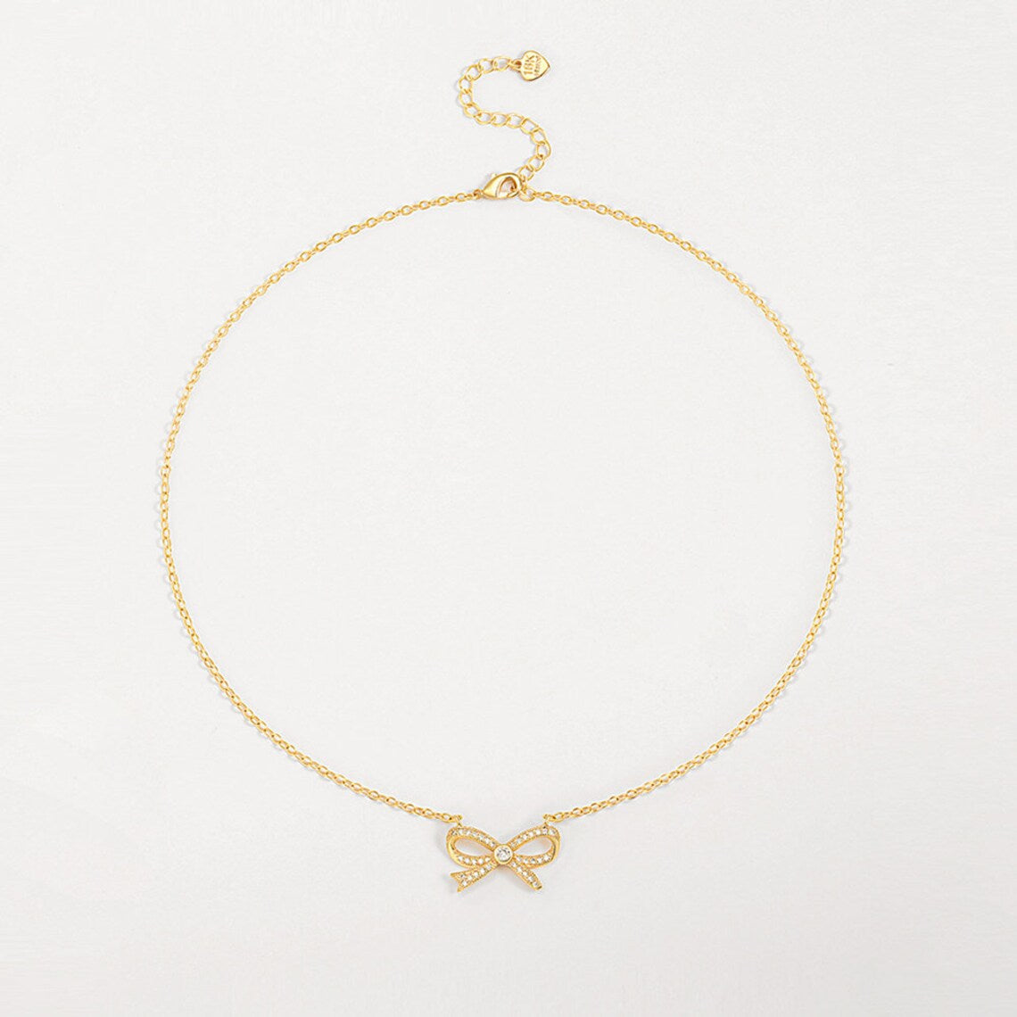 A delicate 18K gold plated cable chain necklace featuring a sparkling cubic zirconia ribbon bow pendant, laid flat against a white surface.