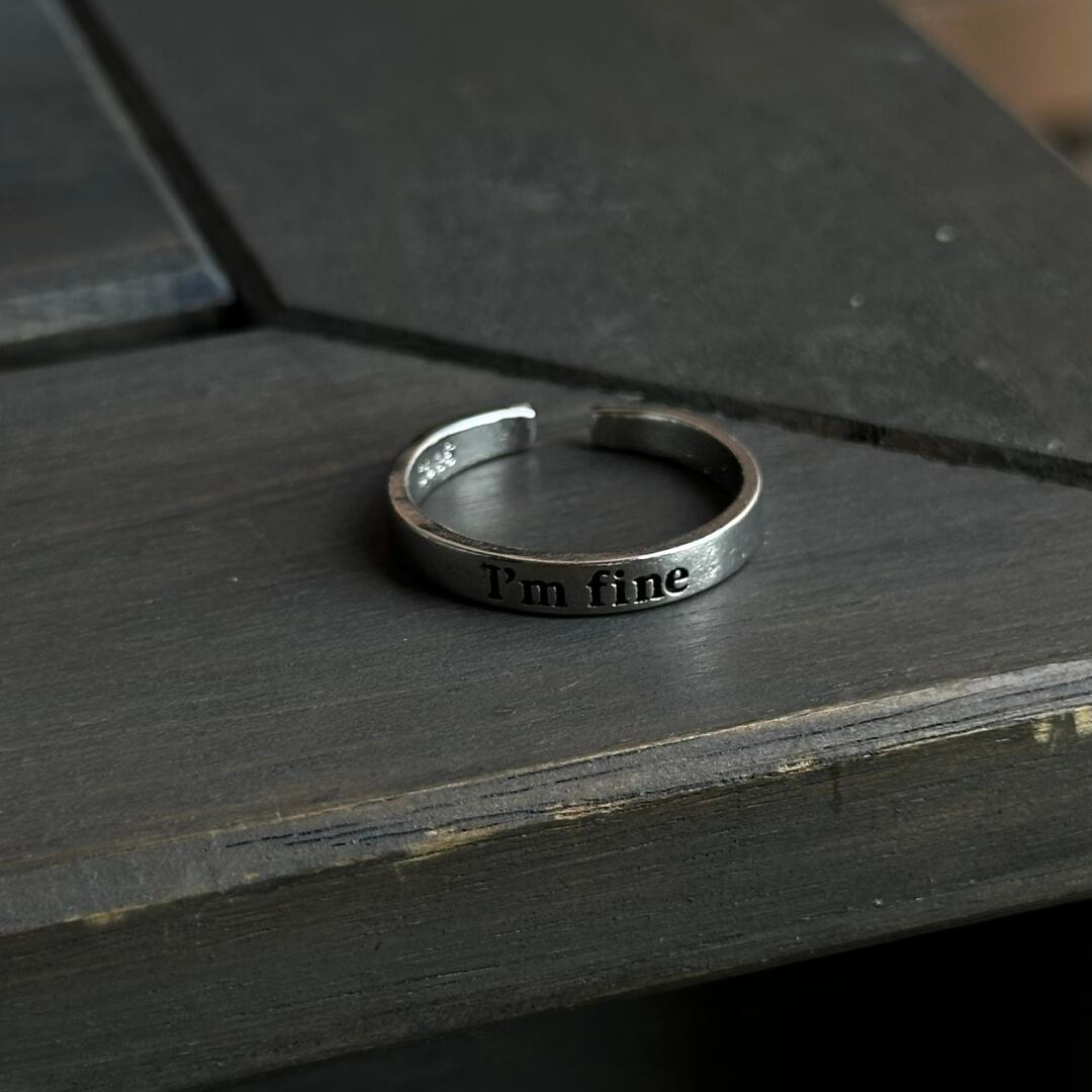 A silver "I'm fine" hidden message ring resting elegantly on a dark, rustic wooden table.