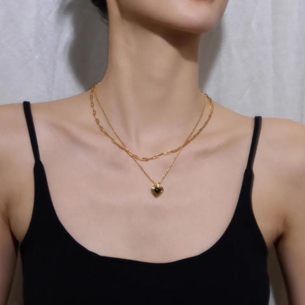 A woman wearing a double-layered black heart necklace with an 18K gold paperclip chain, styled with a simple black tank top.