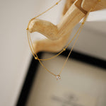 A dainty double-layer gold necklace featuring a beaded satellite chain and a 4mm clear CZ pendant, hanging elegantly from a light wood jewelry display stand.