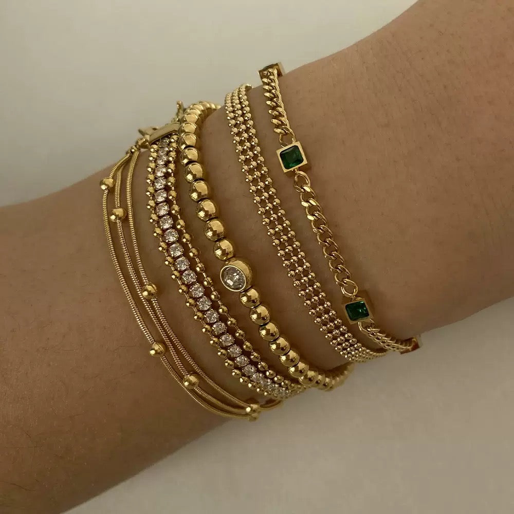 Close-up of a wrist displaying a neat stack of five different gold bracelets, featuring snake chains, a CZ tennis chain, gold beads with a center stone, and a curb chain with square emerald green stones.