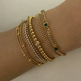 Close-up of a wrist displaying a neat stack of five different gold bracelets, featuring snake chains, a CZ tennis chain, gold beads with a center stone, and a curb chain with square emerald green stones.