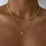 Close-up of a woman's collarbone with a deep tan, wearing a layered 18K gold plated flat snake chain and a sparkling pave initial W pendant.