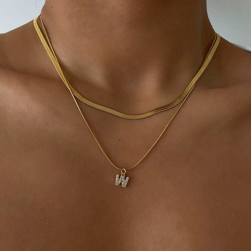 Close-up of a woman's collarbone with a deep tan, wearing a layered 18K gold plated flat snake chain and a sparkling pave initial W pendant.