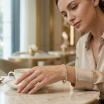 A woman in an elegant beige silk shirt wearing a gold diamond tennis bracelet while enjoying coffee at a cafe.
