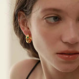 Close-up of a woman wearing chunky 18K gold-plated hoop earrings adorned with colorful star and heart-shaped gemstones.