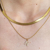 Macro detail shot on a model's neck, clearly showing the woven liquid-like texture of a gold herringbone snake chain paired with a cubic zirconia pave initial H pendant.