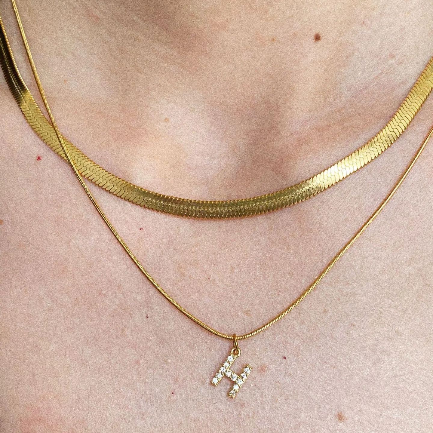 Macro detail shot on a model's neck, clearly showing the woven liquid-like texture of a gold herringbone snake chain paired with a cubic zirconia pave initial H pendant.