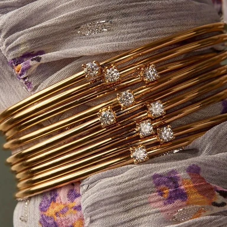 A stack of multiple elegant 18K gold-plated open cuff bangles with small sparkling solitaire diamonds, showcasing a layerable jewelry trend.