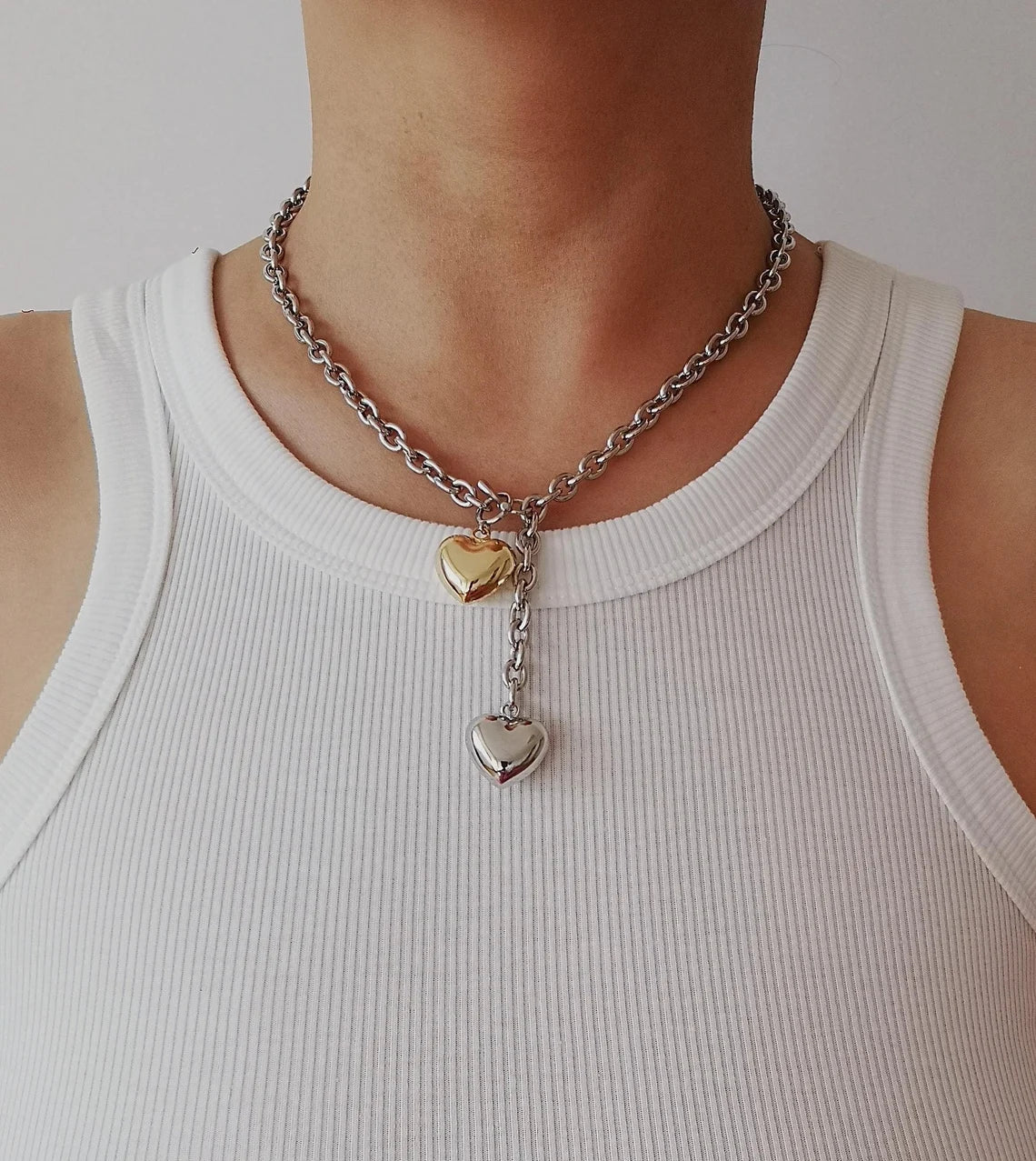 A mixed metal lariat necklace with a chunky silver chain, front toggle clasp, and two dangling puffy hearts in silver and gold, isolated on a white background.