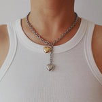 A mixed metal lariat necklace with a chunky silver chain, front toggle clasp, and two dangling puffy hearts in silver and gold, isolated on a white background.