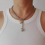 A mixed metal lariat necklace with a chunky silver chain, front toggle clasp, and two dangling puffy hearts in silver and gold, isolated on a white background.