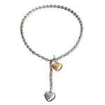 A mixed metal lariat necklace with a chunky silver chain, front toggle clasp, and two dangling puffy hearts in silver and gold, isolated on a white background.