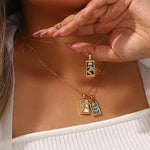 Close-up of a woman wearing a white ribbed top, holding a gold The Magician tarot necklace, beautifully layered with two other tarot pendants, The Lovers and The Empress, resting on her chest.