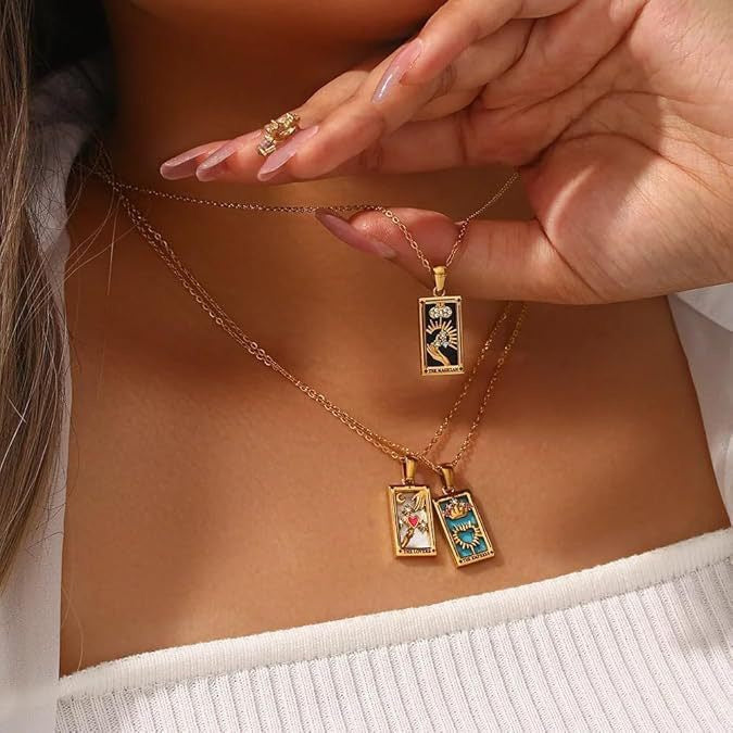 Close-up of a woman wearing a white ribbed top, holding a gold The Magician tarot necklace, beautifully layered with two other tarot pendants, The Lovers and The Empress, resting on her chest.