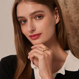 A woman smiling softly with her hand tucked under her chin, wearing a gold angel signet ring and a black and white collared shirt.