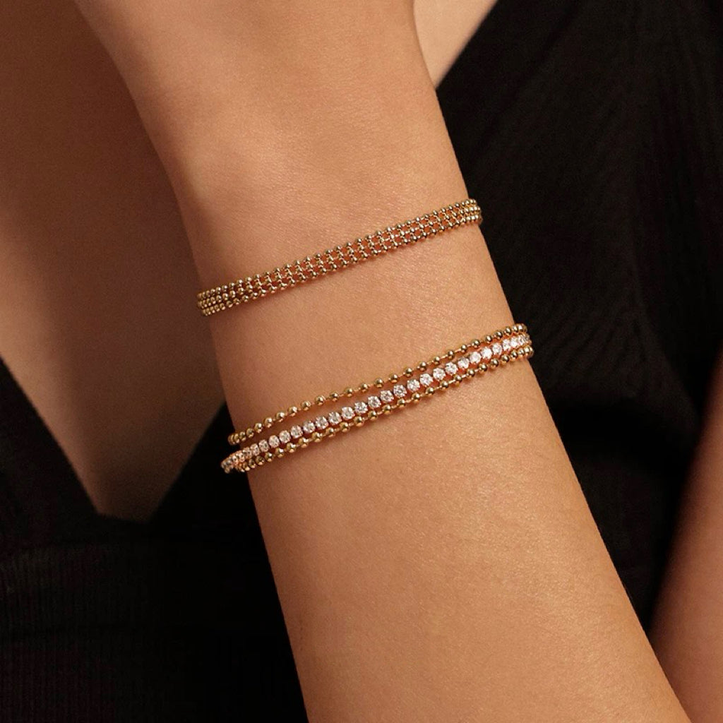 A woman's arm wearing a black top, showcasing a curated wrist stack featuring a triple-row beaded CZ bracelet paired with a thicker gold herringbone chain bracelet.