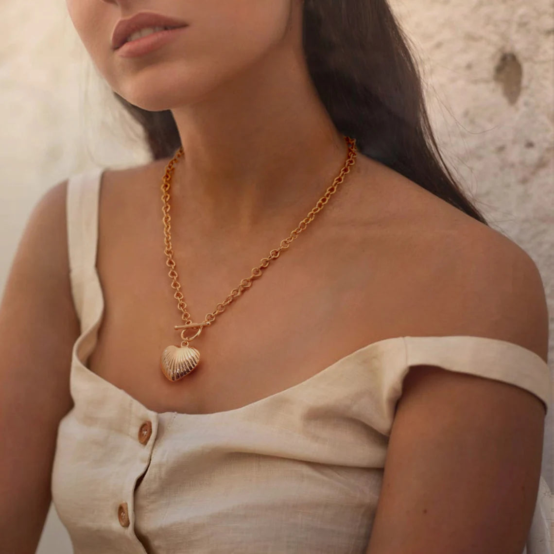 A woman wearing a beige off-the-shoulder top styled with an 18K gold chunky chain necklace featuring a ribbed puffy heart and front toggle clasp.