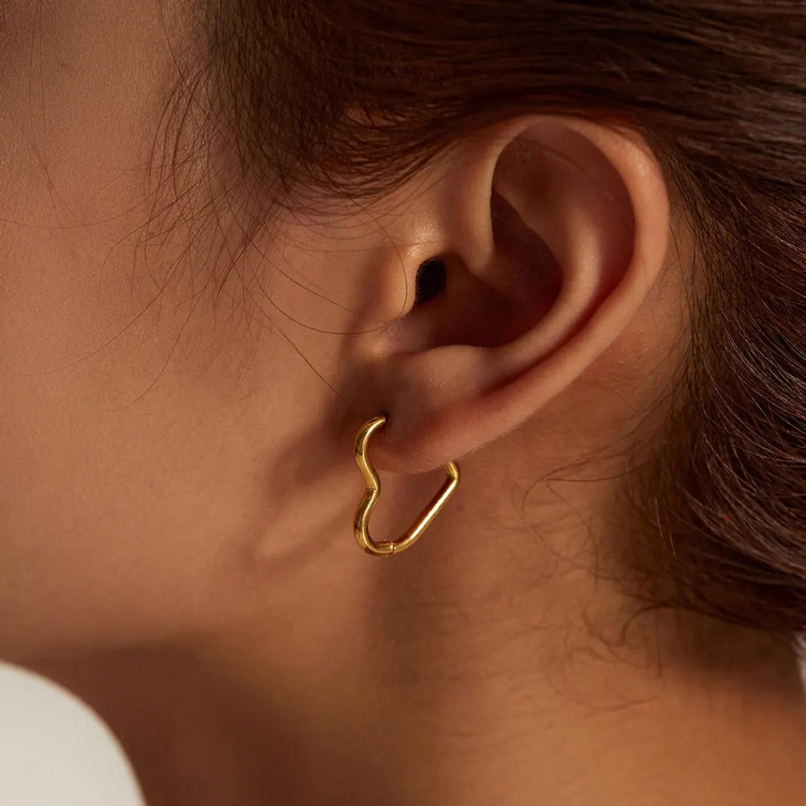 Close-up of a woman wearing a delicate 14K gold plated minimalist heart hoop earring on her earlobe.