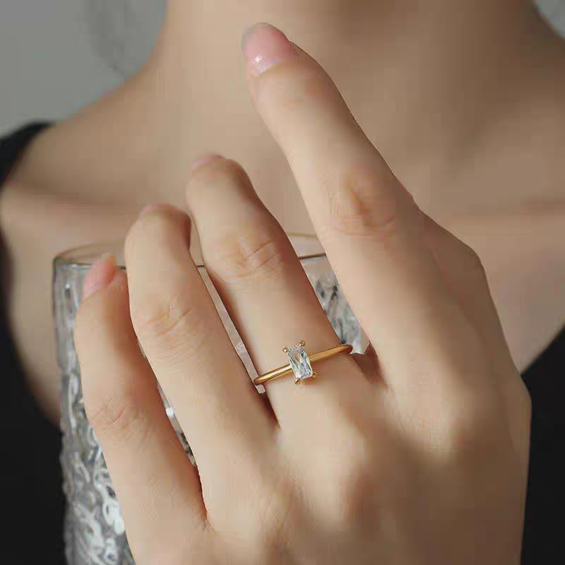 Close-up of a woman's hand holding a textured clear glass, wearing a minimalist gold solitaire ring with a vertically set emerald-cut clear stone.