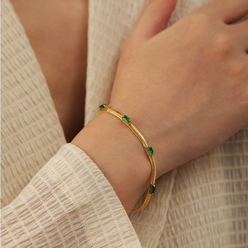 Close-up of a woman wearing a textured beige long-sleeve top with her hand resting on her chest, elegantly modeling a single gold snake chain bracelet with green baguette stones.