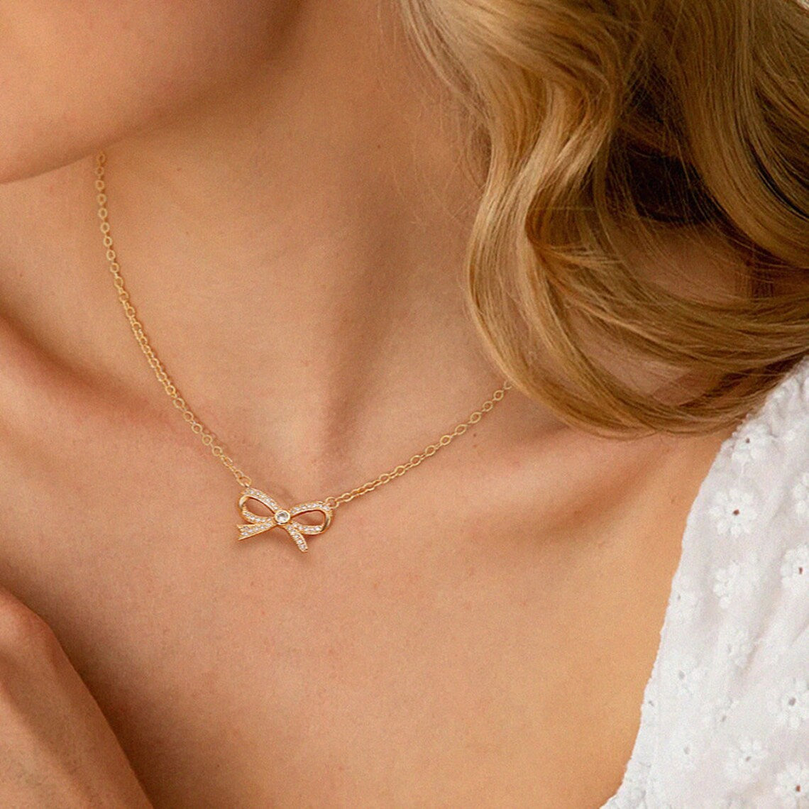A woman wearing a white eyelet top styled with a dainty 18K gold plated pave bow necklace resting elegantly on her collarbone.
