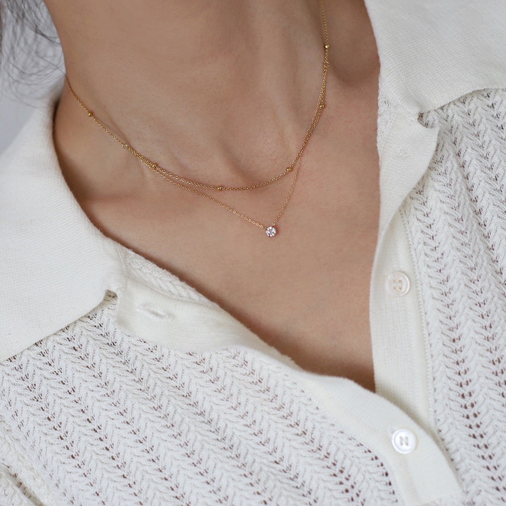 Close-up of a woman's neckline wearing a textured white collared knit shirt, modeling a dainty layered gold necklace featuring a beaded chain and a sparkling solitaire CZ pendant resting on her collarbone.