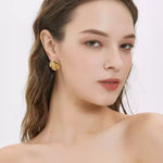 A woman with bare shoulders looking towards the camera, wearing a shiny gold 3D camellia flower stud earring on her earlobe.