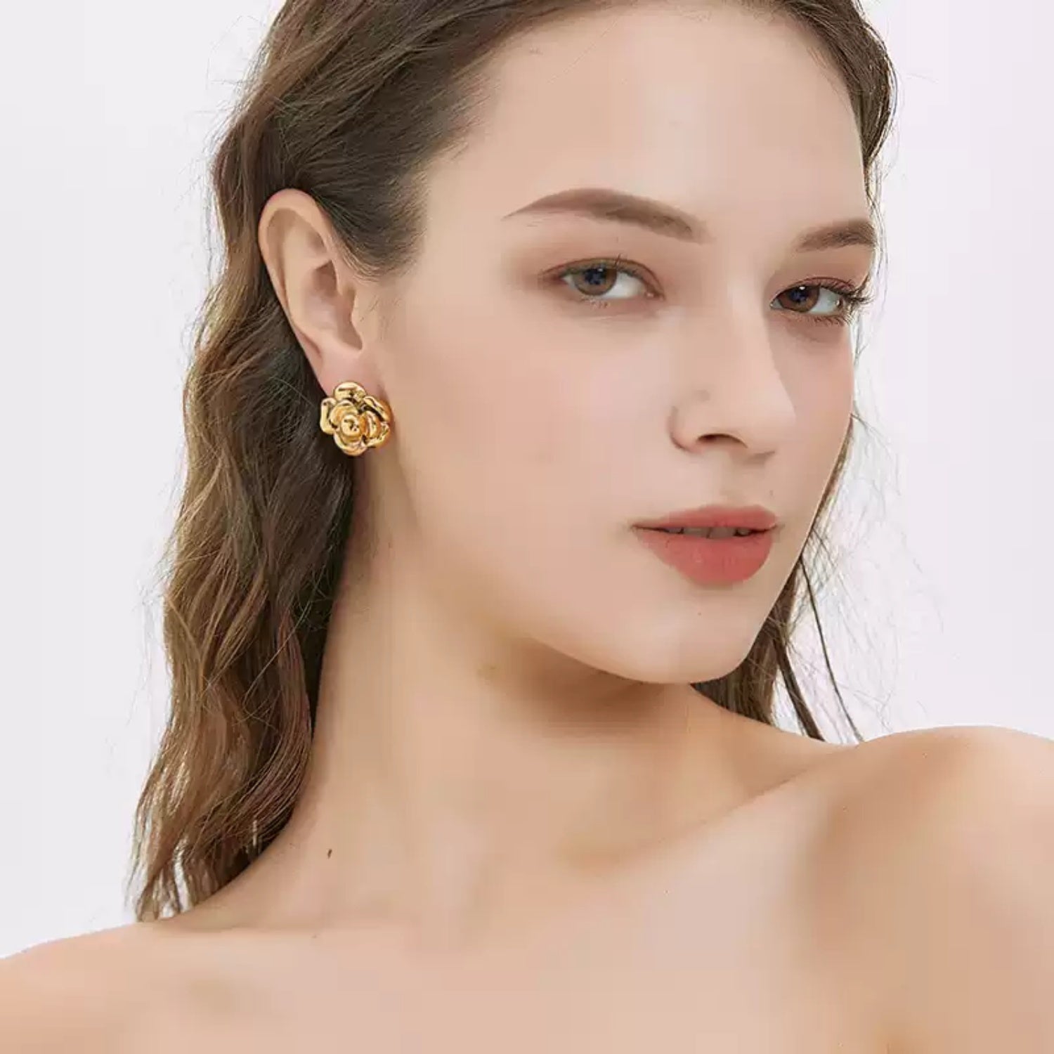 A woman with bare shoulders looking towards the camera, wearing a shiny gold 3D camellia flower stud earring on her earlobe.