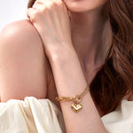 A woman wearing an off-shoulder white top with her hand resting on her opposite shoulder, modeling an 18K gold plated mixed chain puffy heart bracelet.