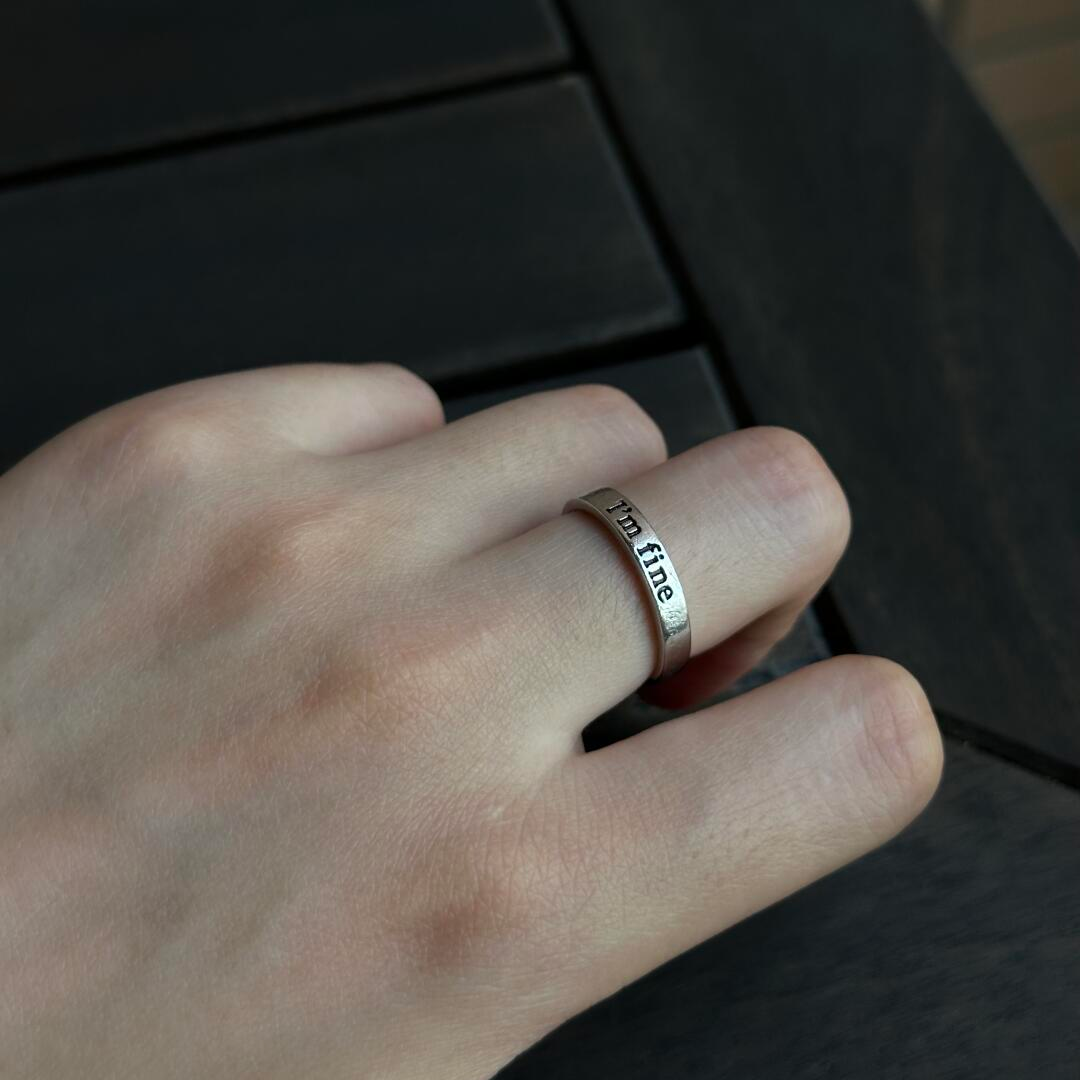 A hand wearing a silver adjustable ring that reads "I'm fine." resting casually on a dark surface.