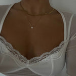 A woman with a deep tan wearing a white sheer lace-trimmed sweetheart top, modeling a layered 18K gold flat snake chain and a pave initial W necklace.