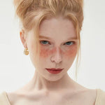 A portrait of a woman with beautiful freckles wearing thick, gold braided hoop earrings featuring a center row of pearls.