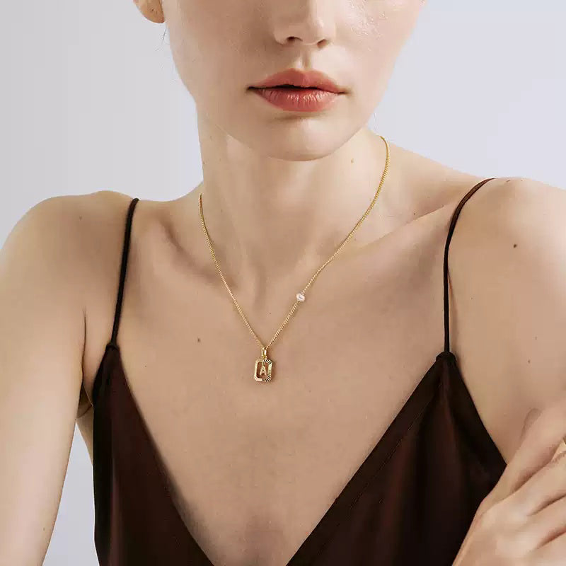 A woman wearing a 14K gold plated vintage sunburst initial necklace with a freshwater pearl, styled with a white top.