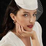 Close-up portrait of a woman with red lipstick and a white birdcage veil resting her hand under her chin, modeling a shiny gold open heart statement earring.