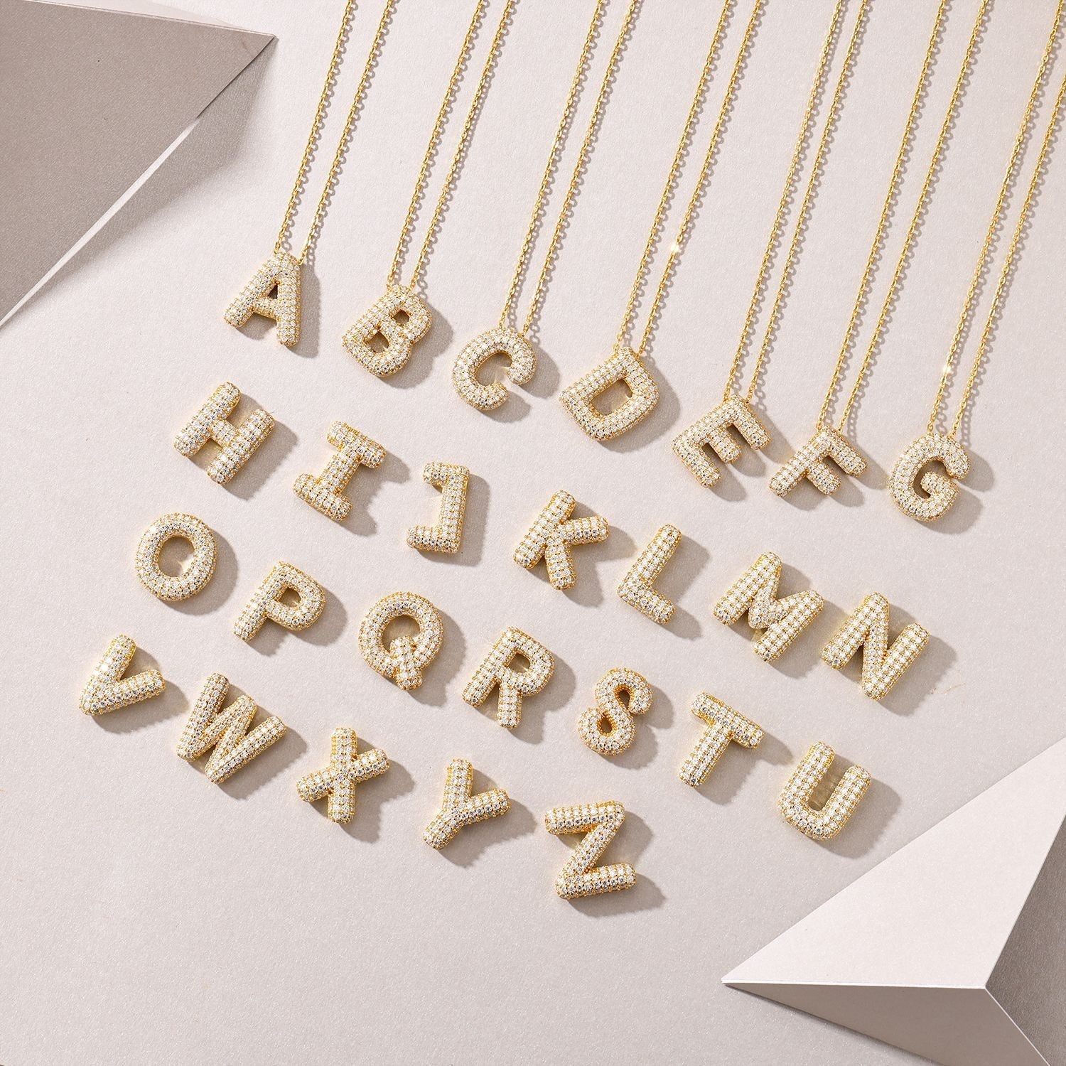 Three 18K gold plated moissanite bubble letter pendants lying flat next to a gold chain on a plain surface.