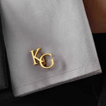 Gold cufflink with 'KG' initials on a gray fabric background